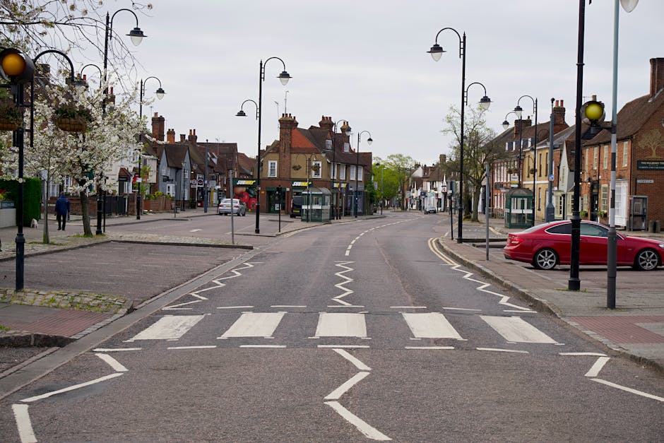 A quiet street scene in a small town with a pedestrian crossing in the foreground, marked by white striped lines on the asphalt. On the left side, there is a paved area with a cherry blossom tree in bloom, surrounded by a low stone border, and a person walking in the distance. The street is lined with traditional lampposts with curved arms, some of which have hanging flower baskets. Several old-style shops and residential buildings, featuring brick and plaster exteriors in shades of beige and brown, are visible along the street, with chimneys on sloped roofs. On the right side, a red car is parked parallel to the curb in a designated parking bay, with curbside pavements and small, green public telephone booths nearby. The background shows the road curving slightly, with more buildings, trees, and overcast skies above, creating a calm, orderly urban environment suitable for showcasing local rubbish and waste collection services.