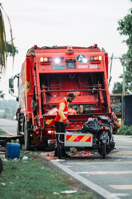 A worker dressed in a bright yellow high-visibility vest and dark trousers is operating a red refuse collection vehicle on a street, with the back of the truck open, revealing mechanical components and hydraulic equipment. The worker is standing near the rear loading area, handling a black garbage bag that is placed on a small platform attached to the vehicle. Nearby, a motorcycle with a black body and silver accents is parked close to the truck, partially obscured by the worker. The scene is set outdoors on a paved street, with a grassy verge and some trees visible to the left, and a building with signage in the background to the right. The sky is overcast, providing diffuse natural lighting that highlights the textures of the vehicle, the worker's clothing, and the surrounding environment. The image reflects a typical street scene involving private waste collection as performed by services like Waste Removal Watford, demonstrating alternative waste handling and rubbish removal in an urban setting.