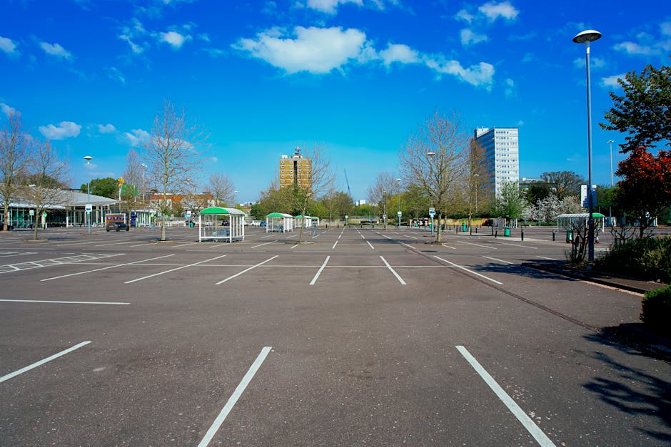 An empty outdoor car park with clearly marked white parking bays on a smooth asphalt surface, situated in an urban area under a bright blue sky with scattered white clouds. The scene features tall, leafless trees along the periphery, along with a few green trees bordering the lot, and several small sheltered structures with green roofs positioned near the edge of the parking area. In the background, multi-storey office or residential buildings are visible, with a mix of modern glass and concrete facades. A tall street lamp stands on the right side of the image, casting a shadow onto the parking surface, while additional street lamps are seen along the perimeter. The environment appears well-maintained with no vehicles parked, and there are no visible signs or markings indicating any restrictions or designated zones. This setting exemplifies a typical private car parking area suitable for access or temporary land use, supporting the context of waste removal and on-site clearance that Waste Removal Watford provides for local businesses or residents performing private waste handling outside of public authority services.