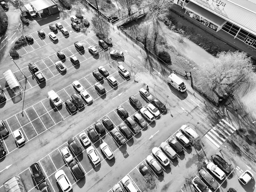 An aerial view of a multi-level parking lot featuring numerous parked cars aligned within clearly marked white parking bays. The vehicles are predominantly small to medium-sized, with a variety of light and dark shades, and the surfaces of the cars are smooth and reflective, indicating newer models or good maintenance. The parking lot surface appears to be asphalt, with some visible wear and faint tire marks. Surrounding the parking area, on the upper right, there is a strip of landscaped greenery with a few trees and small bushes, casting shadows on the ground. A white van is parked near the edge of the lot, adjacent to a pedestrian crossing with white striped markings, suggesting a nearby entrance or exit point. To the upper left, a row of covered bicycle racks is visible, though no bicycles are present. The scene is illuminated by natural daylight, creating shadow patterns that add depth to the layout. This image, associated with waste removal services or alternative rubbish collection, highlights a typical urban parking environment designed for efficient vehicle storage and access, supporting the context of private waste handling or disposal logistics managed by firms like Waste Removal Watford.