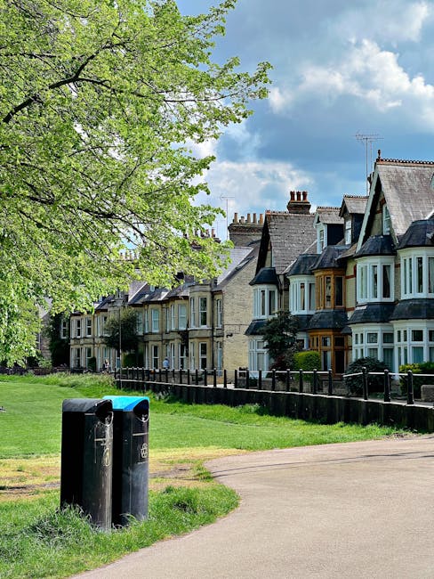 A pair of black waste bins with rounded tops and slightly glossy finishes are situated on a grassy verge next to a paved footpath in front of a row of terraced residential houses. The bins have small slots for waste and are positioned close together, with one slightly behind the other, on a small concrete pad. The background features a line of traditional Victorian-style terraced houses with bay windows, some of which are painted in shades of blue and cream, and brickwork in light brown and beige tones. The houses have pitched roofs with chimney stacks, and some have small front gardens or shrubs in front of their facades. To the left, a large, leafy deciduous tree with bright green foliage extends branches overhead, casting dappled shadow onto the footpath and grass. The sky above is partly cloudy with patches of blue visible between white and grey clouds. The scene appears to be in a quiet suburban area, where private waste collection might be managed independently, supporting rubbish removal services such as those offered by Waste Removal Watford.