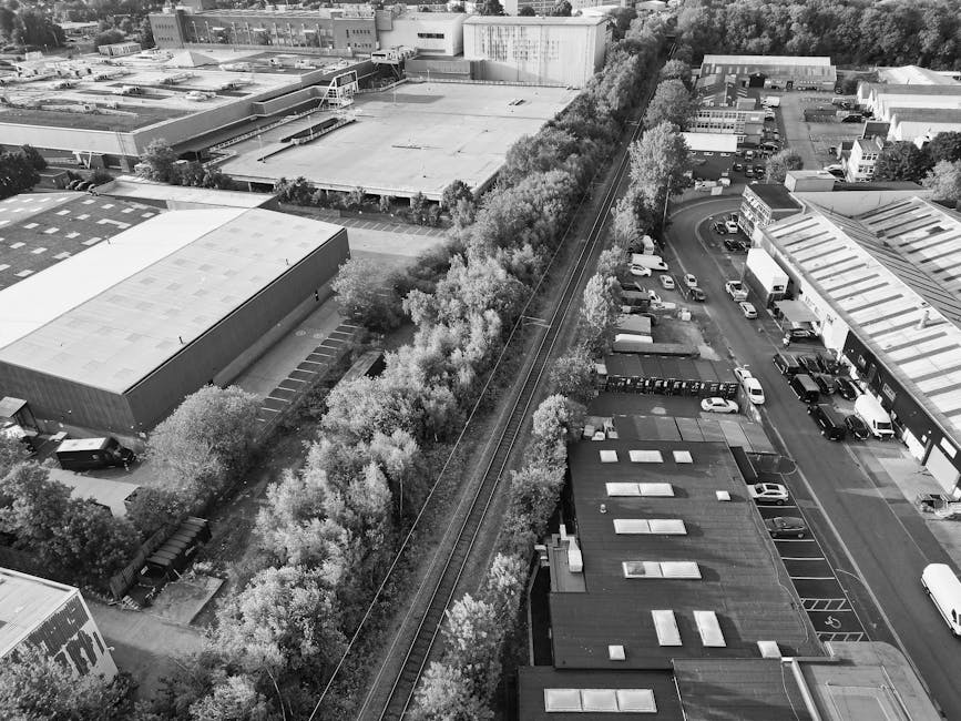 An aerial black-and-white photograph depicts a commercial or industrial area with a railway line running diagonally through the image, flanked by a row of trees with dense foliage on either side. To the left of the railway, there are large warehouse-style buildings with flat roofs, some with visible skylights and vents, and a series of smaller structures nearby. Adjacent to the railway line on the right, there is a parking lot filled with cars, including small and larger vehicles, accessible by a curved road that leads to a cluster of commercial units with metal and glass facades. The surrounding environment features additional industrial or retail buildings with pitched and flat roofs, as well as tree-lined streets and green spaces. This scene relates to private waste management services, as the variety of buildings and parking arrangements suggest scenarios where independent rubbish removal or on-site clearance is involved, aligning with waste removal services available for local commercial properties in the Watford area, such as those offered by Waste Removal Watford.